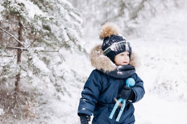 Karlı çam ormanlarında yürüyen bir çocuk. Küçük çocuk kışın dışarıda eğleniyor. Noel tatili. Mavi tulumlu, örgü örüp parkta oynayan tatlı çocuk..