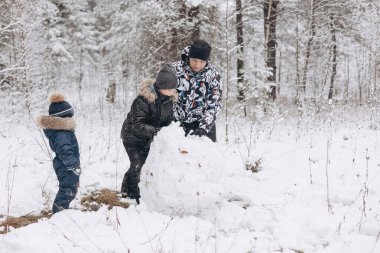 Karlı kış ormanlarında kardan adam için büyük bir kartopu yuvarlayan mutlu baba ve oğullar. Neşeli genç bir adam ve küçük bir çocuk aile yürüyüşünde eğleniyor. Kış zamanı açık hava aktivitesi.