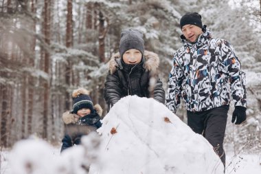 Karlı kış ormanlarında kardan adam için büyük bir kartopu yuvarlayan mutlu baba ve oğullar. Neşeli genç bir adam ve küçük bir çocuk aile yürüyüşünde eğleniyor. Kış zamanı açık hava aktivitesi.