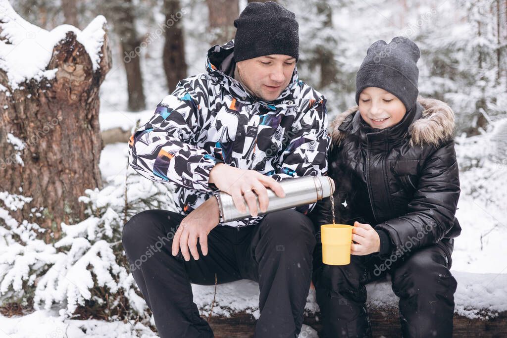 Father and sons drinking tea from thermos and talking sitting together on log in winter snowy forest. Happy man and teenage boys having picnic in winter season outdoors. Local travel. Slow life