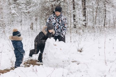 Karlı kış ormanlarında kardan adam için büyük bir kartopu yuvarlayan mutlu baba ve oğullar. Neşeli genç bir adam ve küçük bir çocuk aile yürüyüşünde eğleniyor. Kış zamanı açık hava aktivitesi.