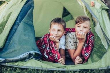 Happy children lying with plaid in camping tent. Family weekend nature outdoor. Smiling teenage boys with mug in hands. Slow life