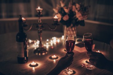 Elleri şarap bardakları tutan adam ve kadın evde mum ışığında romantik bir akşam yemeği yiyorlar. Elleri şarap kadehi tutan adam ve kadın. Sevgililer Günü kavramı mı yoksa mum ışığında gece randevusu mu?.