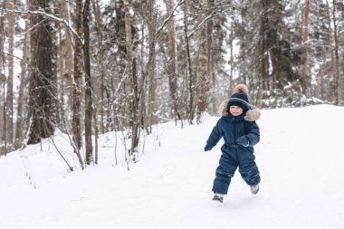 Karlı çam ormanlarında yürüyen bir çocuk. Küçük çocuk kışın dışarıda eğleniyor. Noel tatili. Mavi tulumlu, örgü örüp parkta oynayan sevimli, mutlu bir çocuk..
