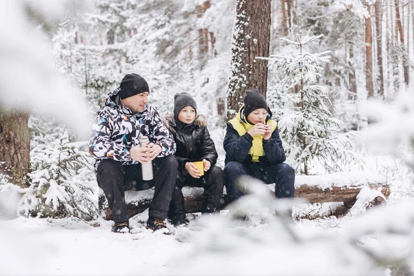 父と息子は魔法瓶からお茶を飲んで 冬の雪の森で丸太の上で一緒に座って話をします 冬の屋外でピクニックをしている幸せな男と10代の男の子 地方旅行 スローライフ — ストック写真