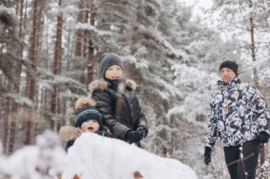 Karlı kış ormanlarında kardan adam için büyük bir kartopu yuvarlayan mutlu baba ve oğullar. Neşeli genç bir adam ve küçük bir çocuk aile yürüyüşünde eğleniyor. Kış zamanı açık hava aktivitesi.