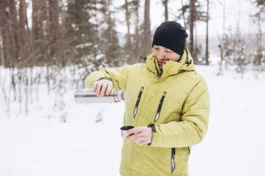 Kışın karlı ormanda bir fincan çay ve termosla gezen genç adam. Beyaz pikniği. Doğada sıcak içecek içmek. Yerel seyahat. Yavaş bir hayat. Kış zamanı açık hava aktivitesi.