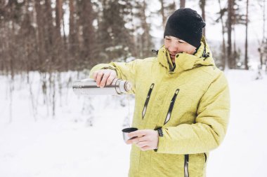 Kışın karlı ormanda bir fincan çay ve termosla gezen genç adam. Beyaz pikniği. Doğada sıcak içecek içmek. Yerel seyahat. Yavaş bir hayat. Kış zamanı açık hava aktivitesi.