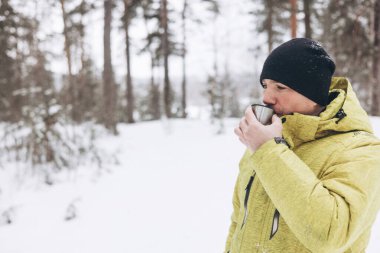 Kışın karlı ormanda bir fincan çay ve termosla gezen genç adam. Beyaz pikniği. Doğada sıcak içecek içmek. Yerel seyahat. Yavaş bir hayat. Kış zamanı açık hava aktivitesi.