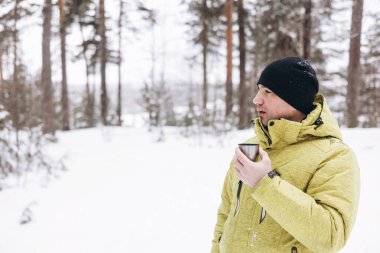 Kışın karlı ormanda bir fincan çay ve termosla gezen genç adam. Beyaz pikniği. Doğada sıcak içecek içmek. Yerel seyahat. Yavaş bir hayat. Kış zamanı açık hava aktivitesi.
