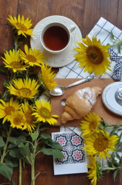 a cup of tea with a croissant on a wooden table surrounded by sunflowers