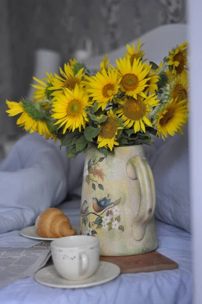 a bouquet of sunflowers in a yellow jug on the bed with a cup of coffee and a plate of croissant and newspaper