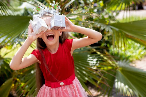 身穿红色衣服的小女孩站在棕榈树旁 用礼品盒闭上她的眼睛 高兴极了 圣诞节 新年在非洲 澳大利亚 喜庆的心情 家庭庆祝活动 — 图库照片