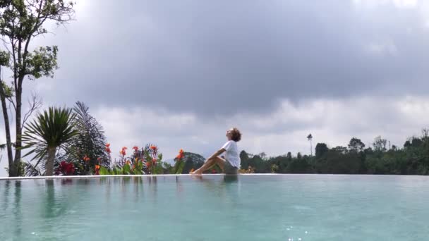 Біла жінка сидить на краю басейну насолоджуючись своїм райським відпочинком — стокове відео