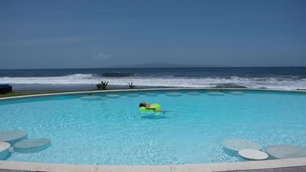 Despreocupado disfrutando en el paraíso, una persona nadando en el anillo inflable amarillo en la piscina azul del hotel cerca de la orilla del mar, tomando el sol y descansando — Vídeos de Stock