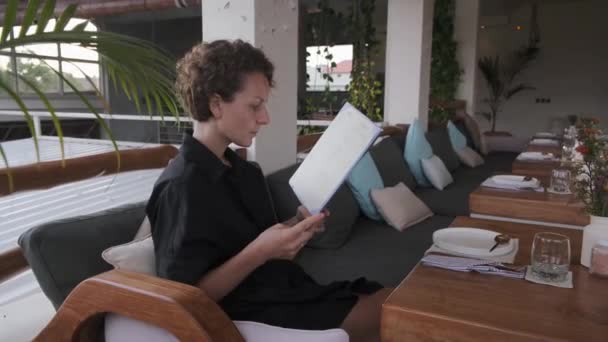 Wanita muda yang santai dengan kemeja hitam yang duduk di restoran di resor mewah, berpikir tentang ketertiban, membaca menu — Stok Video