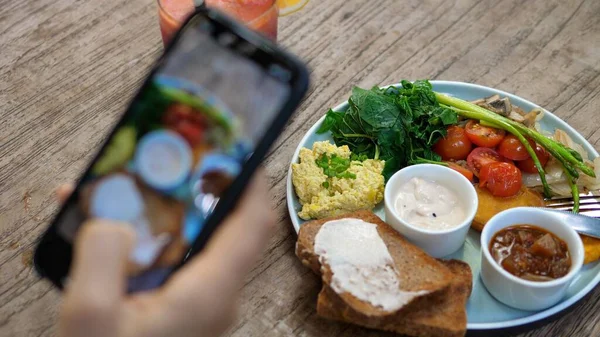 Main d'un blogueur mode de vie sain prend une photo du déjeuner à base de plantes d'une brouille de tofu, des toasts et des légumes servis sur une table en bois — Photo