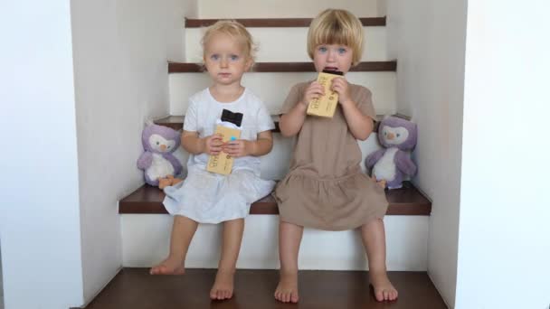 Duas meninas pequenas bonitos em vestidos sentados nas escadas em casa e tentando seus presentes doces da família — Vídeo de Stock