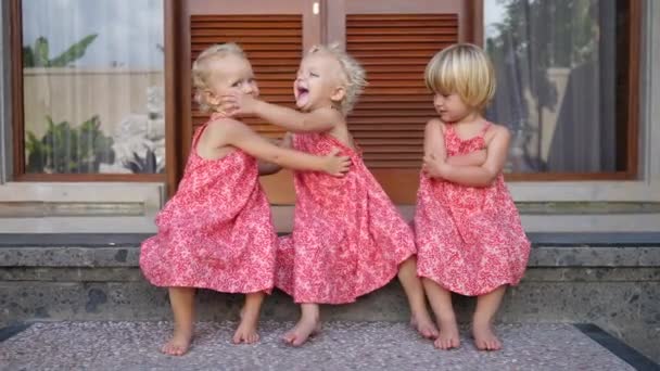 Three adorable blonde toddler girls in pink dresses laughing and cuddling outdoors — стоковое видео