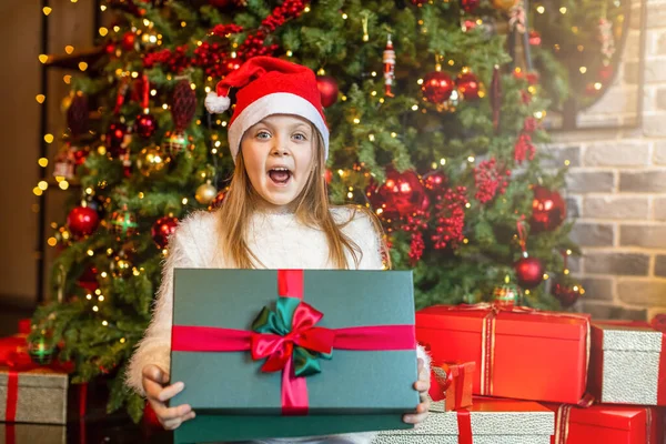 Noel Baba şapkalı mutlu kız Noel ağacı arka planında sihirli bir hediye kutusuna şaşırır. Mutlu Noeller.