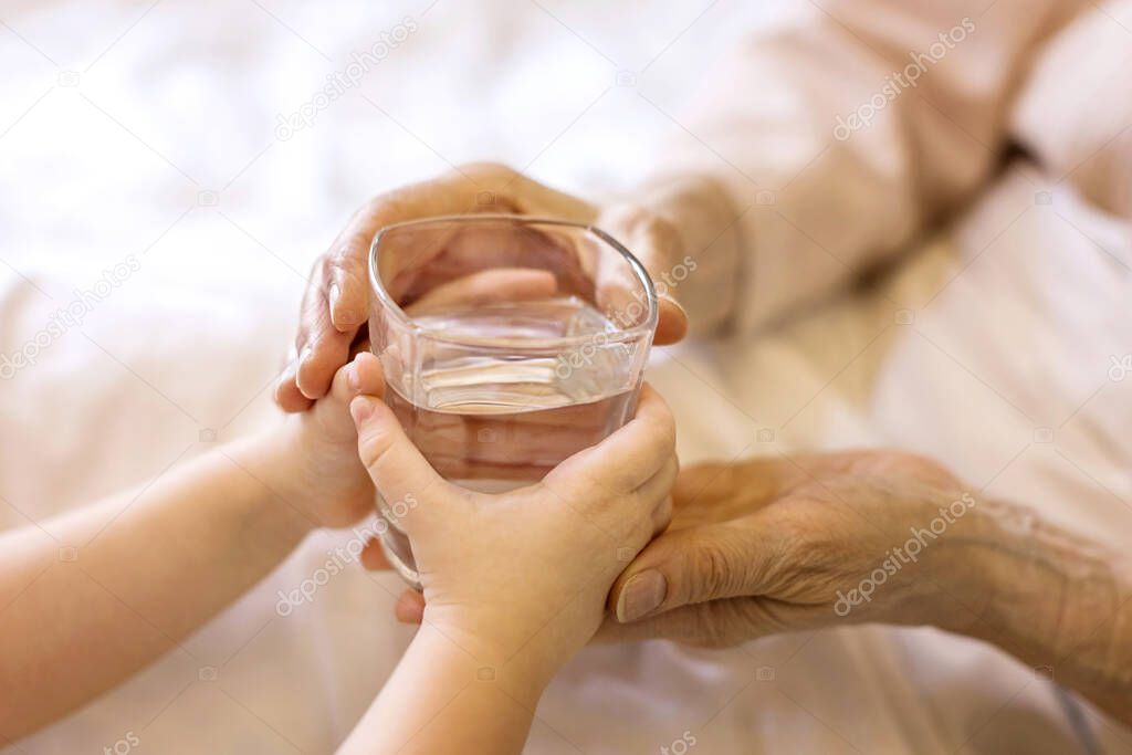 Small child hands gives a glass of water to an old person grandmother in the bed at home. The concept of caring for adults, support, love, assistance, mutual assistance