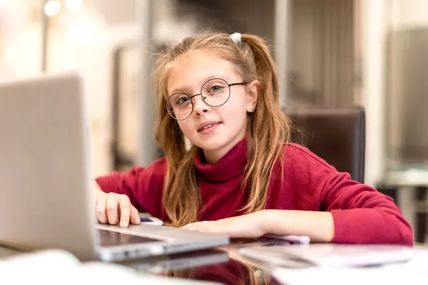 Evde online eğitim ve evde ders çalışmak için dizüstü bilgisayar kullanan mutlu küçük kız evde gülümseyerek kameraya bakıyor. Çocuklar uzaktan öğreniyor. Sosyal uzaklık.