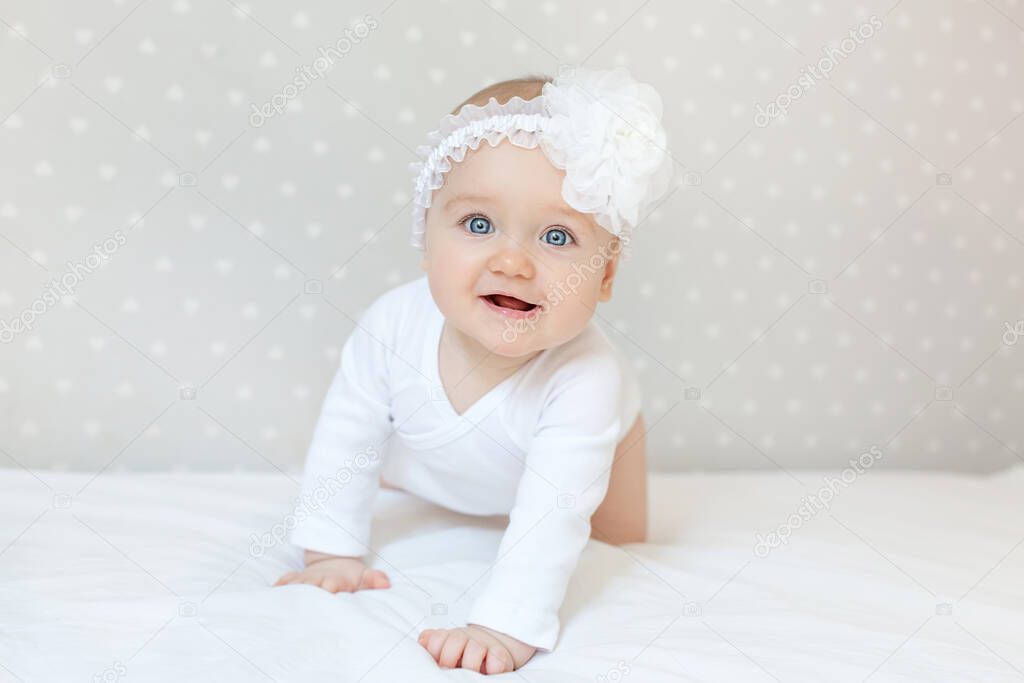 Happy toddler baby girl in a white bodysuit sitting on bed at home and looking at the camera with smile. Copy space for text
