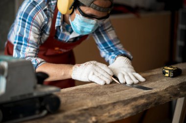 Kahverengi önlüklü genç bir Asyalı marangoz tahta üzerinde çalışırken zımpara makinesi ve mezura kullanıyor. Ahşap fabrikasında çalışan marangoz konsepti. Seçici odak.