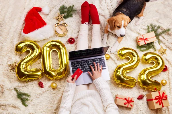 İnternetten alışveriş. Yeni yıl 2022. Bir kız yılbaşı ve yeni yıl için bir dizüstü bilgisayardan hediye alır. Yatakta köpek köpeğinin yanında oturuyordu. Yüksek kalite fotoğraf