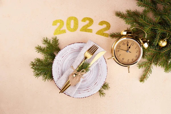 Happy New Year 2022. Festive table setting. Plate, silverware, fir branches, golden alarm clock.
