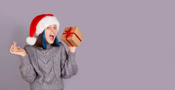 Szczęśliwa dziewczyna w kapeluszu Świętego Mikołaja trzyma prezent z czerwoną wstążką. Sztandar świąteczny, miejsce na reklamę. — Zdjęcie stockowe