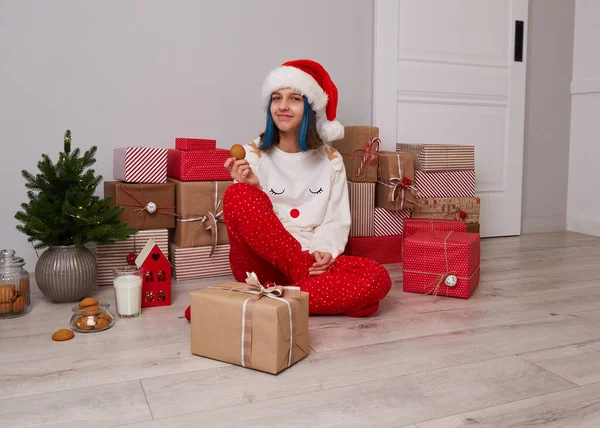 Szczęśliwa dziewczyna w piżamie i kapeluszu Świętego Mikołaja siedzi przy dużym stosie prezentów świątecznych. — Zdjęcie stockowe