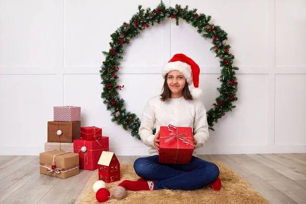 Szczęśliwa dziewczyna w białym swetrze i kapeluszu Świętego Mikołaja otwiera prezent świąteczny — Zdjęcie stockowe