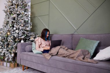 Güzel bir kız, battaniyeyle kaplı bir dizüstü bilgisayarla çalışıyor. Oyuncak ve hediyeli Noel ağacı. Evde rahatlık, Noel tatili..