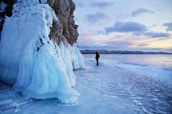 Jezero Bajkal při západu slunce. Na ledě jezera se pohybuje muž. Turista se dívá na rampouchy na skalách. — Stock fotografie