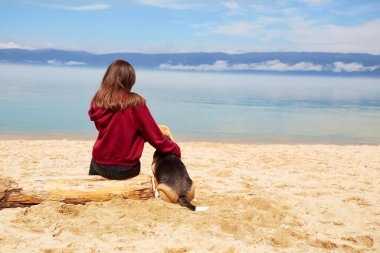 Bir kız kumlu bir sahilde kütüğün üzerinde oturuyor ve köpeğine sarılıyor. Olkhon Adası 'na yaz gezisi, Baykal Gölü. Arkadan bak.. 