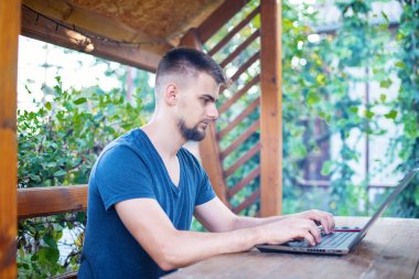 Sakallı genç bir adam dışarıda çalışmak için bilgisayar dizüstü bilgisayar kullanıyor, evde online olarak serbest çalışıyor, yakışıklı bir öğrenci okuyor, öğreniyor, çevrimiçi eğitim alıyor, serbest çalışıyor.