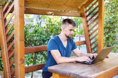 Sakallı genç bir adam dışarıda çalışmak için bilgisayar dizüstü bilgisayar kullanıyor, evde online olarak serbest çalışıyor, yakışıklı bir öğrenci okuyor, öğreniyor, çevrimiçi eğitim alıyor, serbest çalışıyor.