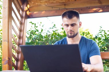 Sakallı genç bir adam dışarıda çalışmak için bilgisayar dizüstü bilgisayar kullanıyor, evde online olarak serbest çalışıyor, yakışıklı bir öğrenci okuyor, öğreniyor, çevrimiçi eğitim alıyor, serbest çalışıyor.