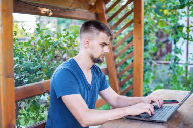 Sakallı genç bir adam dışarıda çalışmak için bilgisayar dizüstü bilgisayar kullanıyor, evde online olarak serbest çalışıyor, yakışıklı bir öğrenci okuyor, öğreniyor, çevrimiçi eğitim alıyor, serbest çalışıyor.