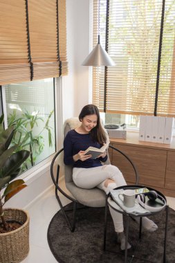 Ev konseptinden güzel bir kadın modern bir sandalyede oturmuş dekore edilmiş minimum bir odada ünlü bir kitap okuyor..