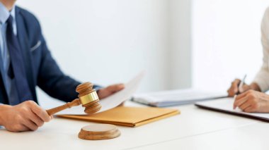 Hukuk konsepti. Derin mavi takım elbisedeki hukuk temsilcisi, yardımcısı onunla aynı masada otururken şirketin ona sunduğu resmi belgeyi onaylamak için tokmak kullanıyor..