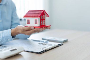 Emlakçının müşterisine sunduğu emlak konsepti kırmızı ev modelini avuçlarına koyup ellerini biraz kaldırarak....