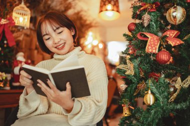 Noel ağacının önünde genç bir kadın evde Noel süsleriyle kitap okuyor.