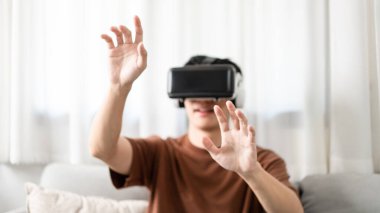 Teknoloji kavramı. Kahverengi tişörtü ve açık mavi kotlu adam koltukta otururken sanal gerçeklik kulaklığı takıyor..