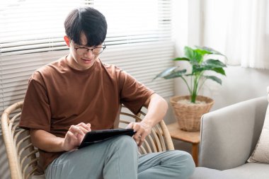 Teknoloji kavramı. Günlük tişörtü ve kotuyla rahat bir şekilde tahta koltukta oturan ve iPad 'deki web tarayıcısını kontrol etmek için dokunmatik ekran yapan erkek..