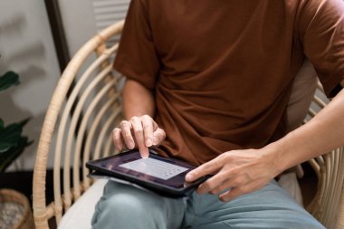 Teknoloji kavramı. Günlük tişörtü ve kotuyla rahat bir şekilde tahta koltukta oturan ve iPad 'deki web tarayıcısını kontrol etmek için dokunmatik ekran yapan erkek..