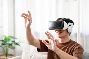 Teknoloji kavramı. Kahverengi tişörtü ve açık mavi kotlu adam koltukta otururken sanal gerçeklik kulaklığı takıyor..