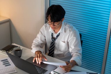 Fazla mesai. Ofis personeli masasında oturmuş, evrak okuyor ve not defterine yazı yazıyor..