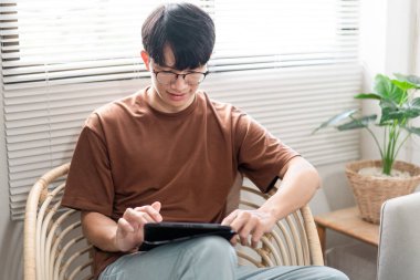 Teknoloji kavramı. Günlük tişörtü ve kotuyla rahat bir şekilde tahta koltukta oturan ve iPad 'deki web tarayıcısını kontrol etmek için dokunmatik ekran yapan erkek..